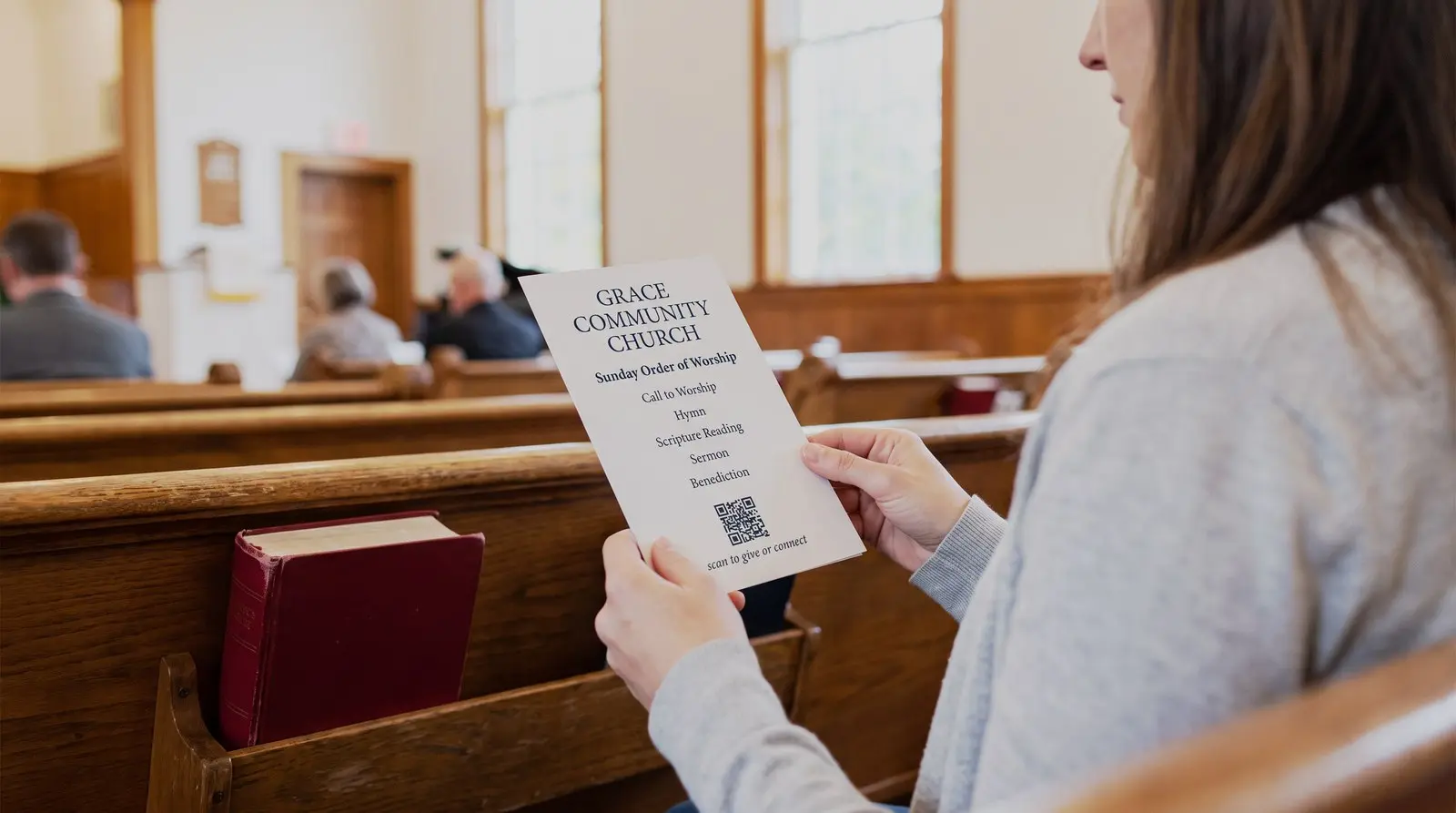 A woman seated in a wooden pew holding a Sunday bulletin with a QR code captioned scan to give or connect, a red hymnal on the pew shelf and the congregation softly blurred beyond