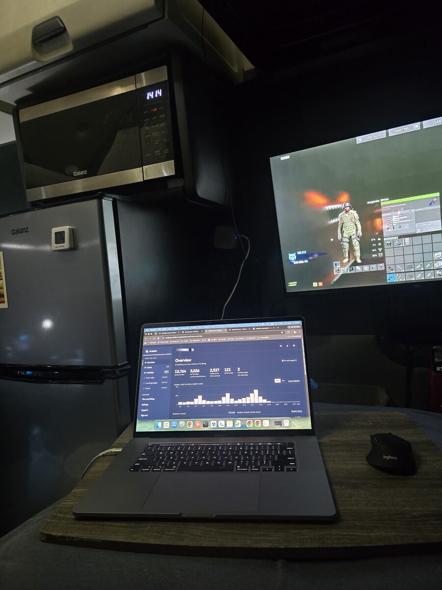 Inside a truck sleeper at night: a MacBook on a wooden lap desk showing the Graklink dashboard, a TV playing a Rust video, a microwave and mini fridge stacked behind.